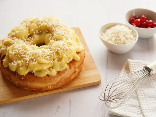 Rosca de Pascua decorada con crema pastelera y azúcar granulada, lista para disfrutar. A su lado, cuencos con cerezas y ralladura, y un batidor.