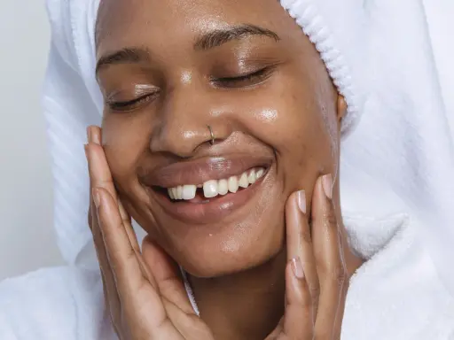 Visage radieux d'une femme souriante, peau hydratée après l'utilisation de rétinol. Elle porte une serviette sur la tête et un peignoir blanc.