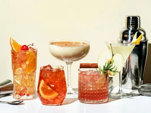 Eine Auswahl an fruchtigen Cocktails, garniert mit Orangen, Äpfeln und Kirschen, neben einem Cocktail-Shaker.