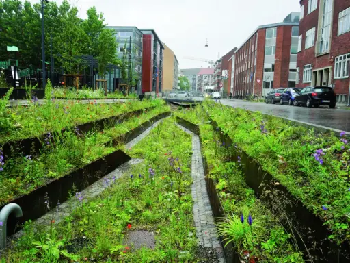 Grüne Oase mit blauer Infrastruktur: Ein Stadtgarten mit Wegen aus Kopfsteinpflaster und metallischen Beeteinfassungen, umgeben von üppiger Vegetation und Blumen.