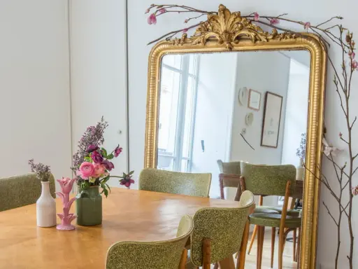 Un grand miroir ancien doré, orné de branches fleuries, reflète une salle à manger avec des chaises vertes. Quelle couleur peindre un miroir ancien ? L'or reste un classique intemporel.