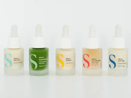 Cinq flacons de sérum et crème hydratante aux couleurs vives, chacun avec une étiquette indiquant son usage : anti-soif, imperfections, unifiant, anti-grisaille, anti-âge.