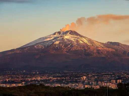 Etna fumante, con la cima innevata che brilla al tramonto, domina una città siciliana.