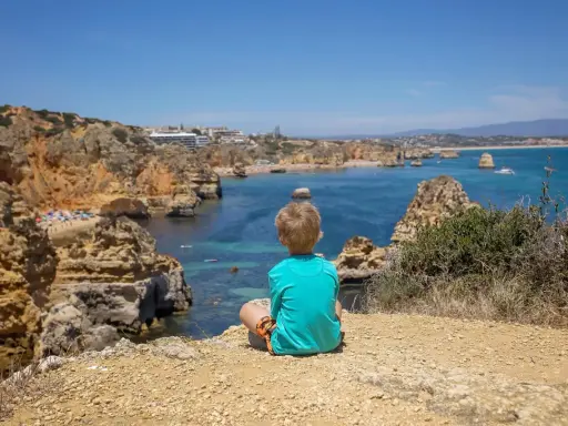 Kleinkind blickt auf die Küste Portugals. Felsen, türkisfarbenes Meer und eine Bucht mit Strand.