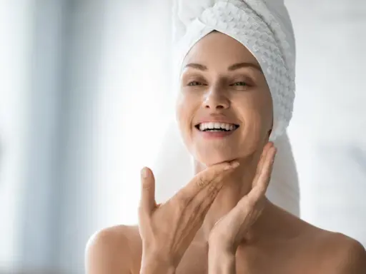Une femme souriante, serviette sur la tête, applique délicatement de l'eau micellaire sur son visage pour une peau fraîche et nette.