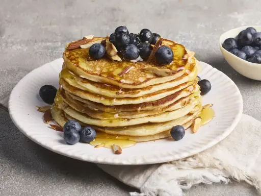 Stapel fluffiger High Protein Pancakes mit Blaubeeren und Mandelsplittern, beträufelt mit Honig.