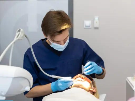 Dentysta w niebieskim fartuchu i masce wykonuje zabieg. Pacjentka ma założone okulary ochronne. Nowoczesne znieczulenie komputerowe zapewnia komfort.