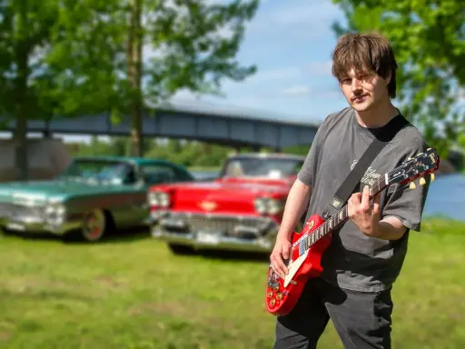 Erik Meduza spelar gitarr framför klassiska bilar vid en sjö.