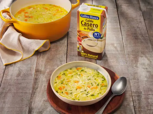 Un tazón humeante de sopa de pollo casera, con fideos finos y trozos de zanahoria, junto a una olla amarilla y un cartón de caldo de pollo.