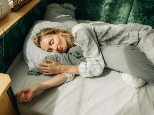 En kvinna sover lugnt i sängen, omfamnande en kudde. Solstrålar faller in genom fönstret och skapar en känsla av guidad avslappning.