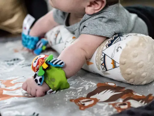 Rączka 6-miesięcznego dziecka z kolorową zabawką na nadgarstku, leżącego na macie z motywem zwierząt.