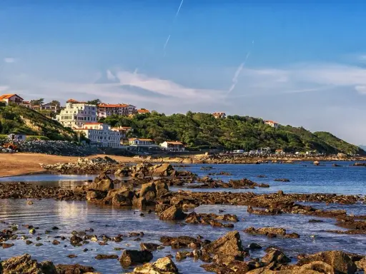 Vue côtière avec des maisons blanches sur une colline verdoyante, une plage de sable et des rochers dans l'eau. Le cadastre zone inondable est visible.
