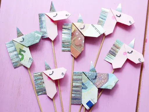 Kreatives Geld falten: Mehrere Einhorn-Figuren aus Geldscheinen und Glitzerpapier auf Holzstäbchen vor rosafarbenem Hintergrund.