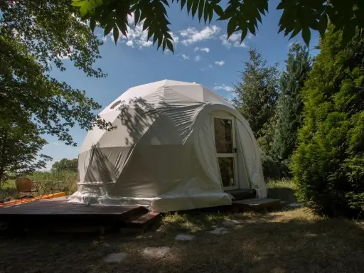 Biały namiot kopułowy, idealny na glamping noclegi, otoczony zielenią. Drewniany taras z meblami zachęca do relaksu.