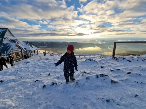 Radosne bieszczady z dziećmi. Mała dziewczynka w czerwonej czapce cieszy się śniegiem na szczycie góry, w tle widać schronisko i piękne zachmurzone niebo.