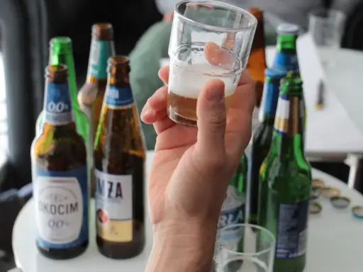 Ręka trzyma kieliszek z piwem bezalkoholowym. W tle kilka butelek piwa, idealne na przerwę w pracy.