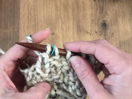 Manos tejiendo con agujas de madera un punto de relieve con lana gruesa y un marcador azul.