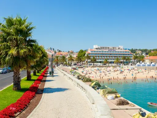 Sonniger Strandtag in Cascais bei Lissabon. Palmen säumen die Promenade, Menschen genießen das Meer und das Hotel Baía ragt empor.