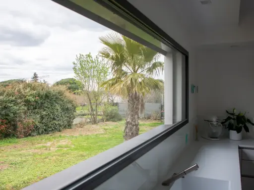 Vista desde una cocina moderna con **ventanas automáticas** que enmarcan un jardín con palmera y vegetación frondosa.