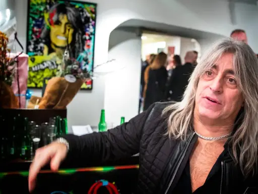 Mikkey Dee son, en man med långt grått hår och en silverfärgad kedja, poserar vid ett bord med blommor och flaskor.