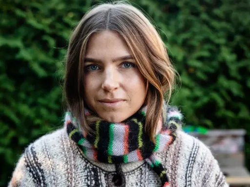Elin Berlin med blå ögon och fräknar, klädd i en stickad tröja och randig halsduk, står framför en grön häck.
