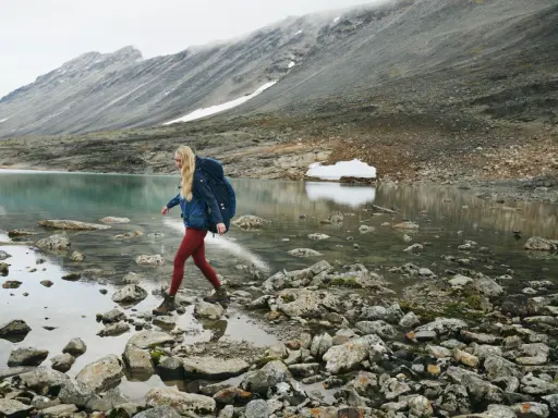 Kobieta z plecakiem przemierza kamieniste brzegi górskiego jeziora. W tle skaliste zbocza, idealne dla skandynawskie marki outdoorowe.