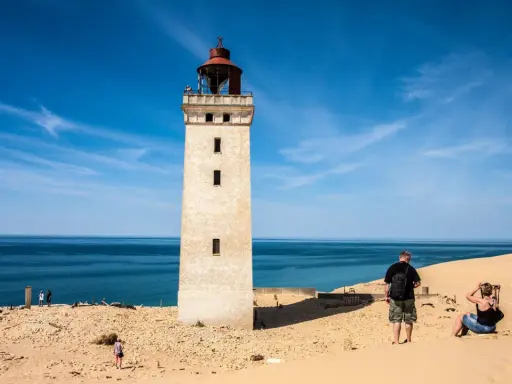 Ett vitt utsiktstorn i Danmark reser sig mot en blå himmel. Sanddyner och hav omger tornet.