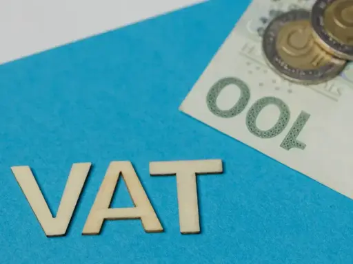 Drewniane litery VAT na niebieskim tle, obok banknot 100 zł i monety. Symbolizuje zwolnienie z VAT.