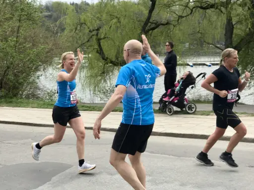 Två löpare ger varandra high-five under roliga lopp. En kvinna i blått och en man i blått. I bakgrunden syns en barnvagn och en sjö.