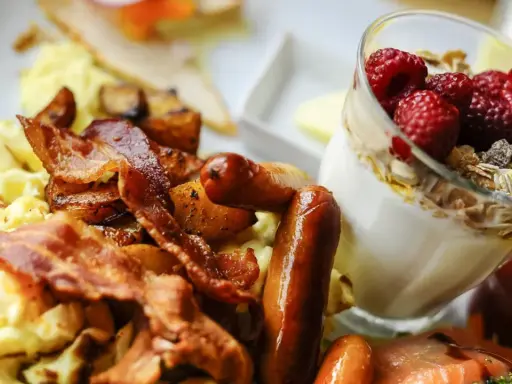 Herzhafte Ideen zum Brunch: Rührei, Speck, Würstchen und Kartoffeln neben einem Glas Joghurt mit Beeren und Müsli.