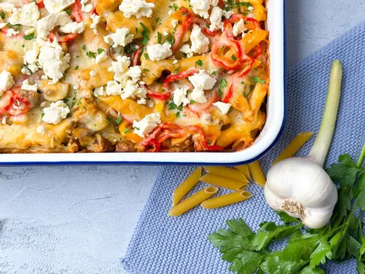 Herzhafter Gyros Auflauf mit Penne, Paprika und Feta, garniert mit Petersilie. Daneben liegen Penne, Knoblauch und frische Kräuter.