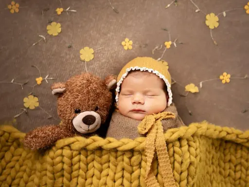 Bambino addormentato con orsacchiotto, avvolto in una coperta gialla. Un'idea per foto neonati fai da te con decorazioni floreali.