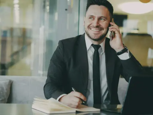 Uśmiechnięty mężczyzna w garniturze rozmawia przez telefon, notując coś w zeszycie. To właśnie taki mikroprzedsiębiorca buduje swój biznes.