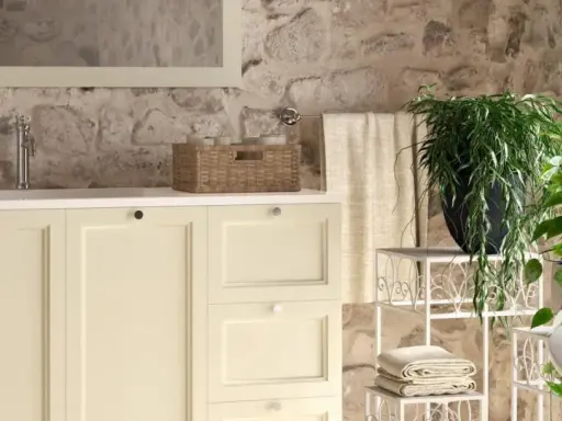 Bagni rustici di campagna con mobile lavabo color crema, cesto in vimini, piante verdi e pareti in pietra grezza.