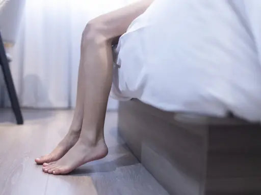 En person sitter på sängkanten, benen dinglar. Kanske söker hen en restless legs huskur för att finna ro.