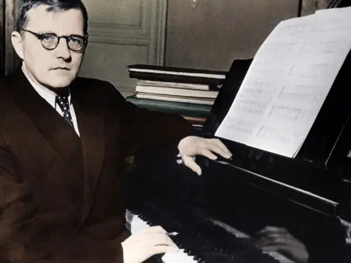 Dmitri Szostakowicz w okularach siedzi przy fortepianie, grając z nut.