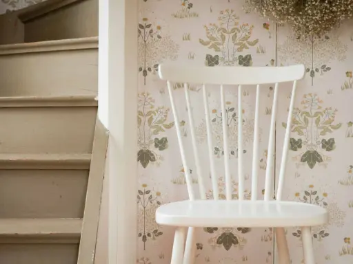 En vit stol står framför en blommig tapet. Bredvid syns en trappa som väntar på att måla om bord och stolar.
