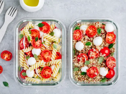 Zwei Glasbehälter mit frischen Mahlzeiten: links Nudelsalat, rechts ein bunter Quinoa-Salat mit Tomaten und Mozzarella.