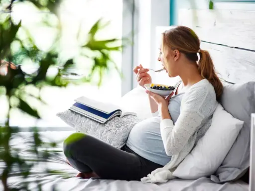 Gravid kvinna med svullen mage äter frukost i sängen. En bok ligger bredvid.