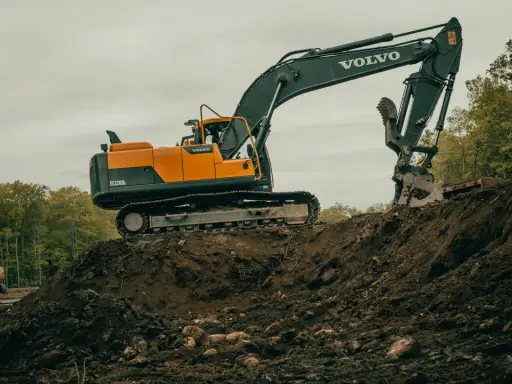 Pomarańczowo-czarna koparka Volvo na placu budowy, z innym mniejszym sprzętem w tle.