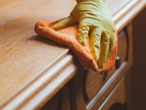 Main d'une personne portant un gant jaune, nettoyant un meuble en bois ciré encrassé avec un chiffon orange.