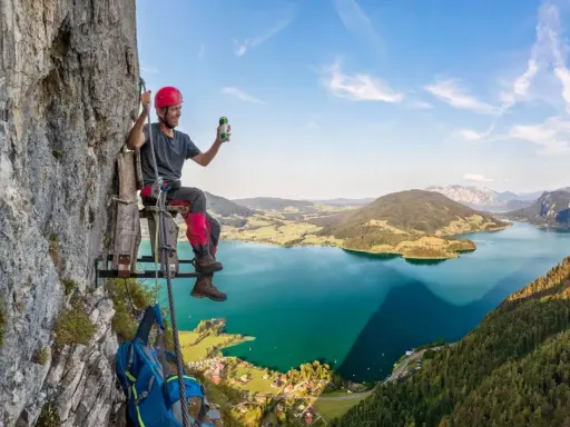 Mężczyzna w kasku siedzi na drewnianej platformie przy skalnej ścianie, ciesząc się widokiem na jezioro w Austrii. To wspaniała przygoda na via ferrata Austria!