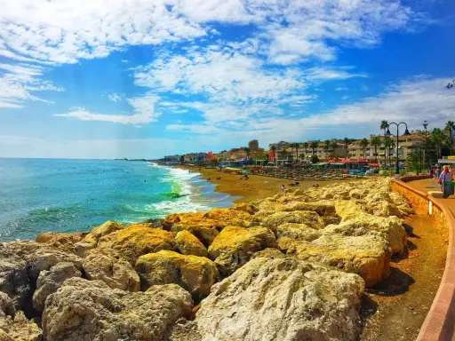 Sonniger Tag an der playa de la carihuela. Türkises Meer, goldener Sandstrand mit Liegen und Palmen, gesäumt von bunten Gebäuden. Felsen im Vordergrund.