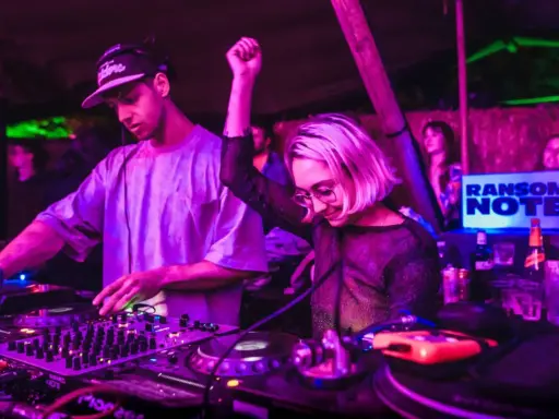 DJ set b2b: un uomo e una donna mixano musica in un locale con luci viola e verdi. La folla balla.