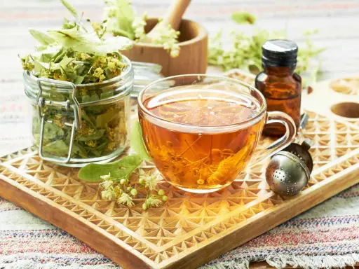 Tee aus Lindenblüten: welcher tee bei heiserkeit hilft, ist hier als dampfendes Getränk in einer Tasse, daneben getrocknete Blüten und ein Tropfenfläschchen.