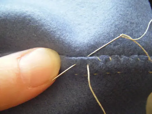 En tumme håller tyg medan en nål med tråd syr för hand olika stygn.