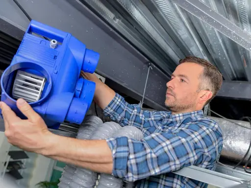 Un technicien installe un boîtier bleu pour une installation VMC double flux, connectant des conduits flexibles sous un plafond métallique.