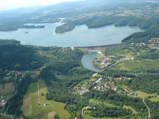 Widok z lotu ptaka na zapora wodna w Myczkowcach, otoczona zielonymi wzgórzami i rozległym jeziorem.