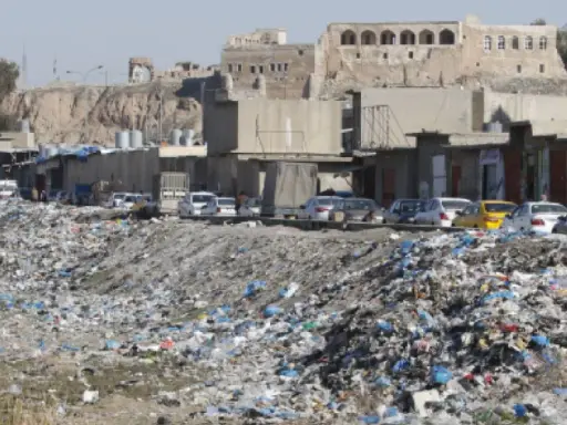 Müllberge säumen die Straßen von Kirkuk, Irak. Autos parken hinter den Geschäften, während im Hintergrund alte Gebäude auf einem Hügel stehen.