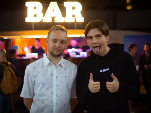 Dwóch mężczyzn pozuje przed napisem "BAR". Jeden w koszuli w palmy, drugi w bluzie z napisem SBM, pokazuje kciuki w górę i wystawia język.